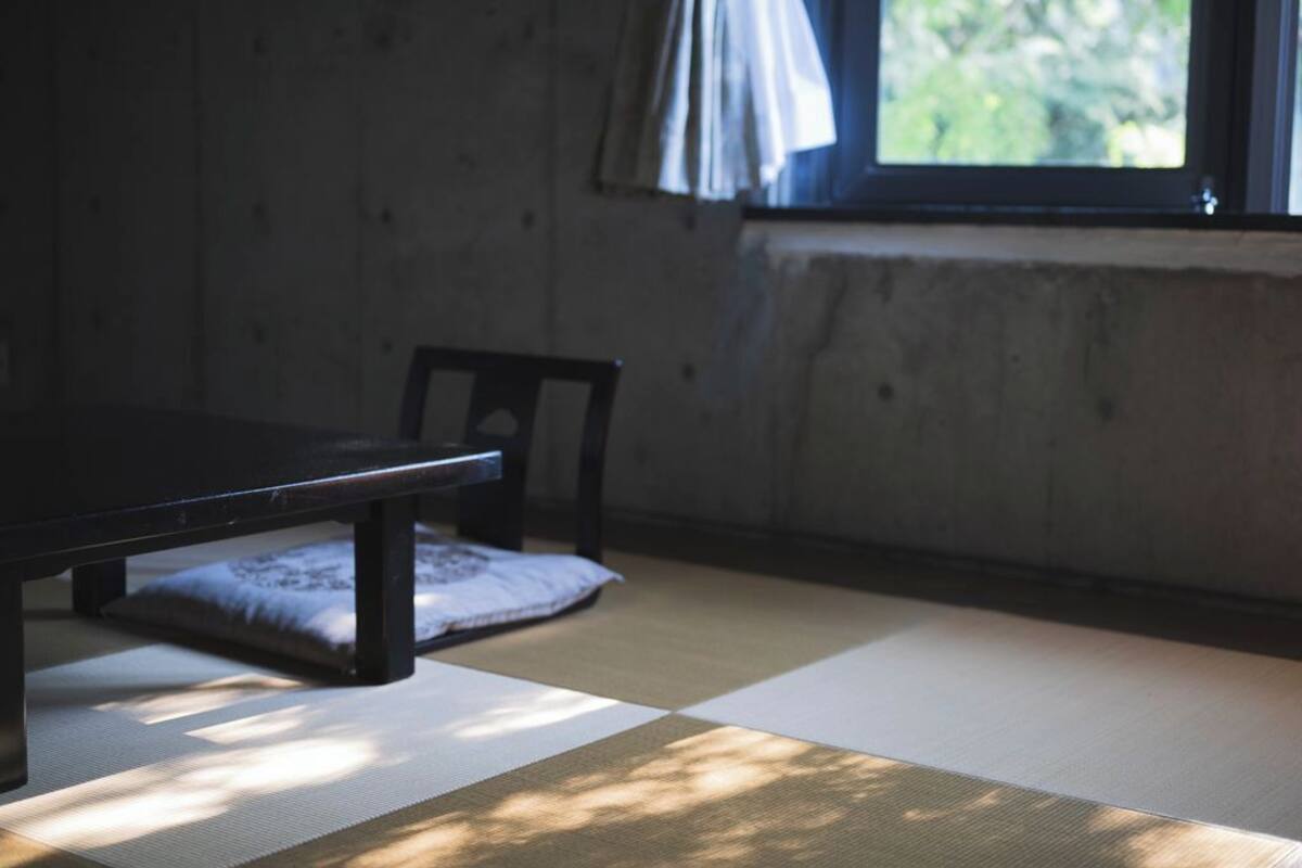 Tatami-mattoilla päällystetty huone, jossa on matala pöytä ja tuoli, ikkuna verhoilla sekä näkymä vehreään maisemaan.