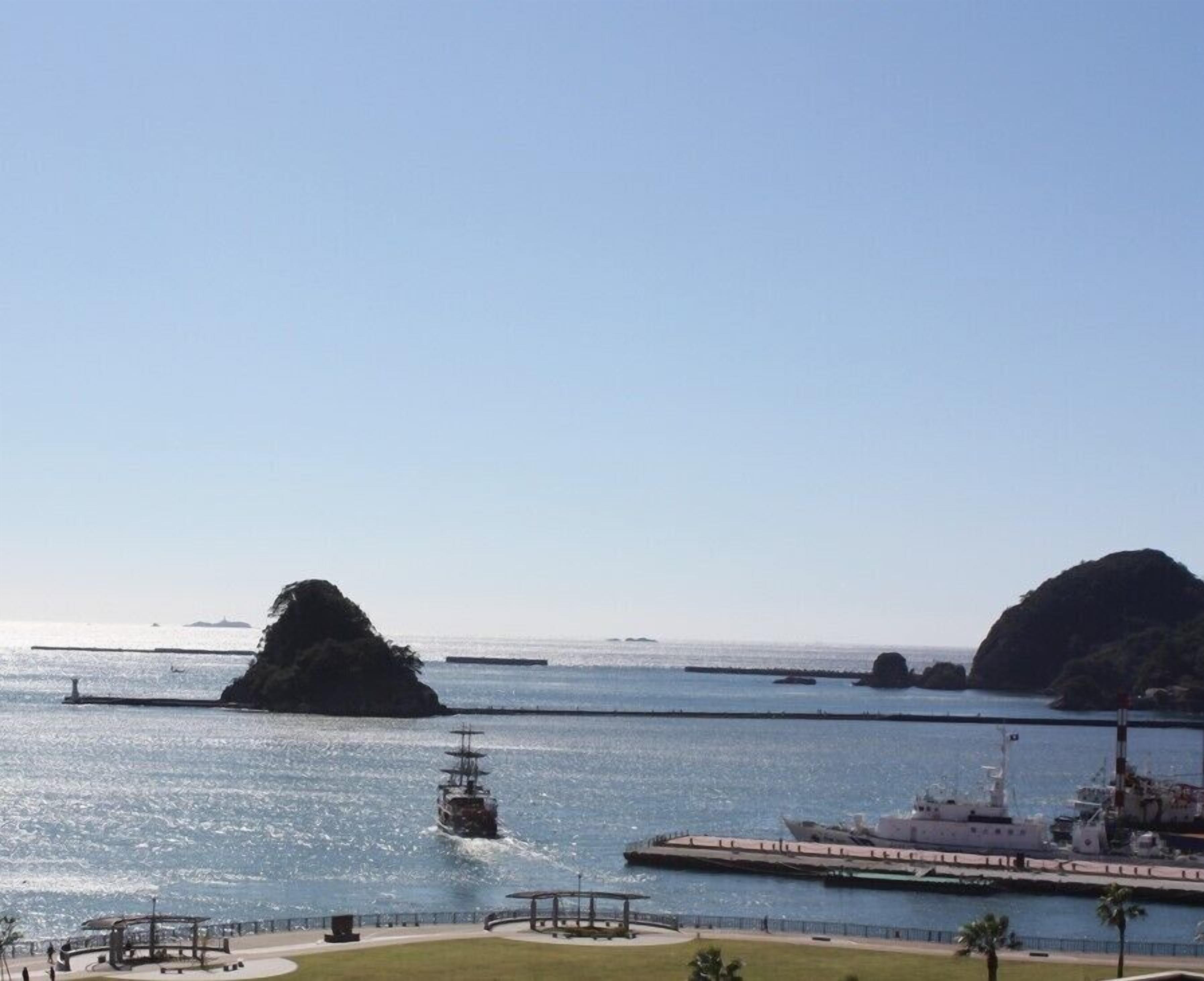 A coastal view with a large ship docked at a pier, a smaller ship in the water, and a small island in the distance.