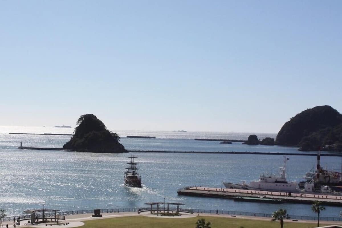 A coastal view with a large ship docked at a pier, a smaller ship in the water, and a small island in the distance.