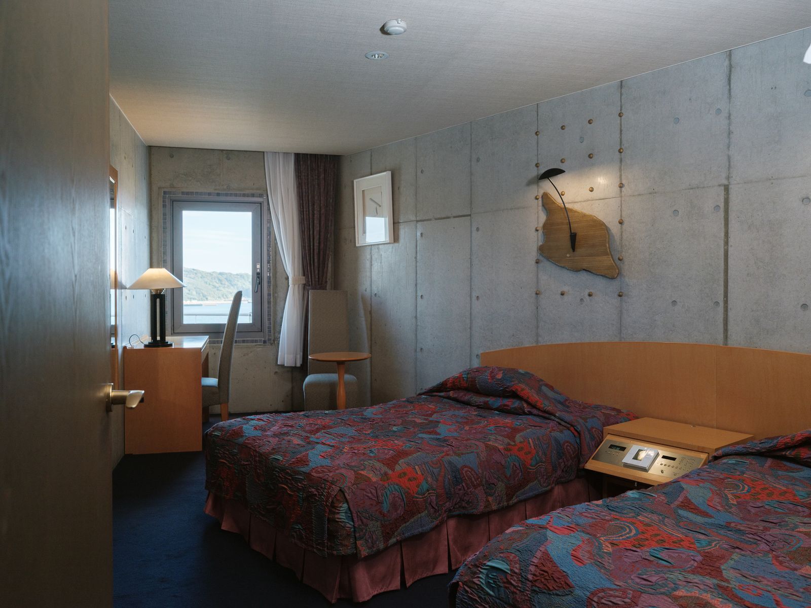 A hotel room with two beds, a desk, a chair, a window with curtains, and a wall-mounted light fixture.