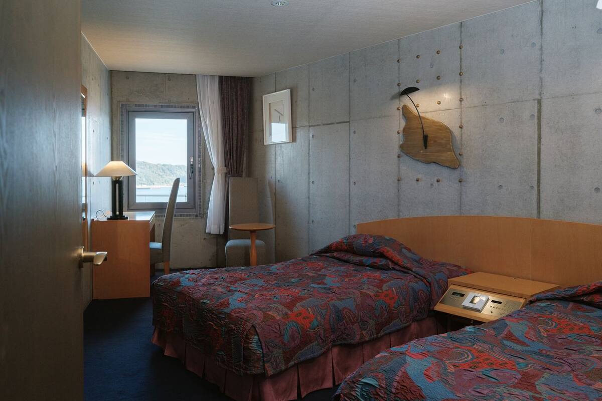 Hotellihuone, jossa on kaksi sänkyä, työpöytä, tuoli, ikkuna verhoilla ja seinään kiinnitetty valaisin.