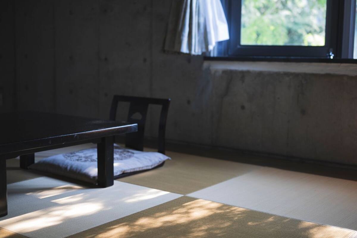Tatami-mattoilla päällystetty huone, jossa on matala pöytä ja tuoli, ikkuna verhoilla sekä näkymä vehreään maisemaan.