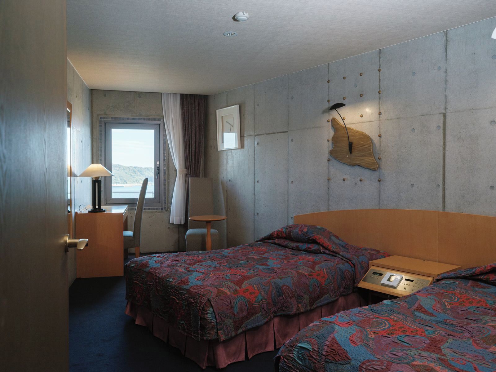 A hotel room with two beds, a desk, a chair, a lamp, and a window with a view.