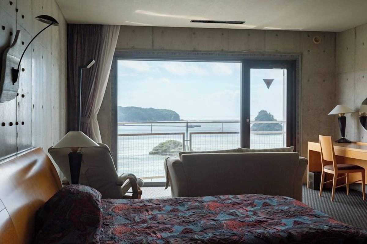 Ett hotellrum med en säng, ett skrivbord, en stol, ett fönster med utsikt över havet och en balkong.