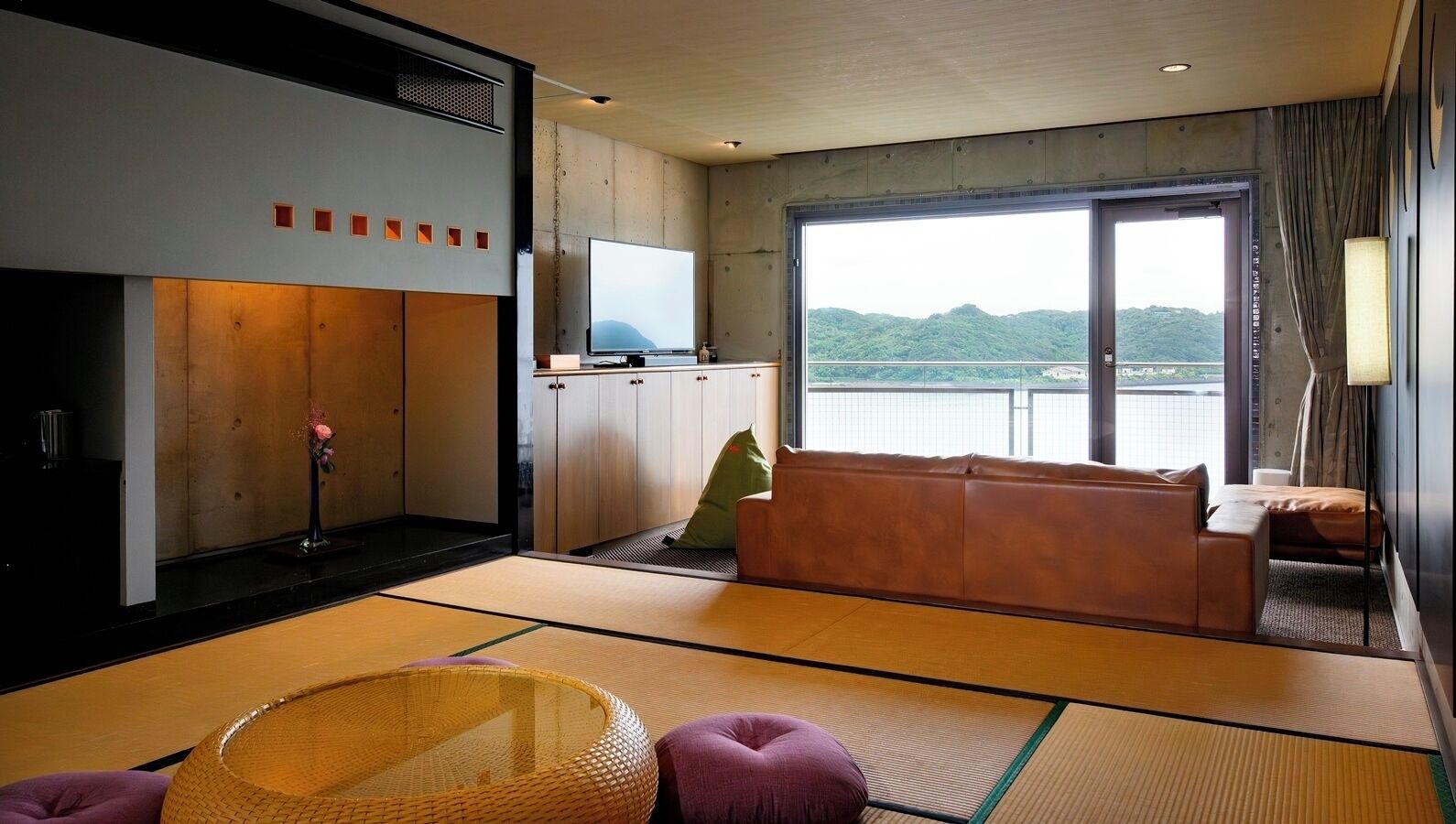 A room with tatami flooring, a sofa, a television, and a view of a mountain.