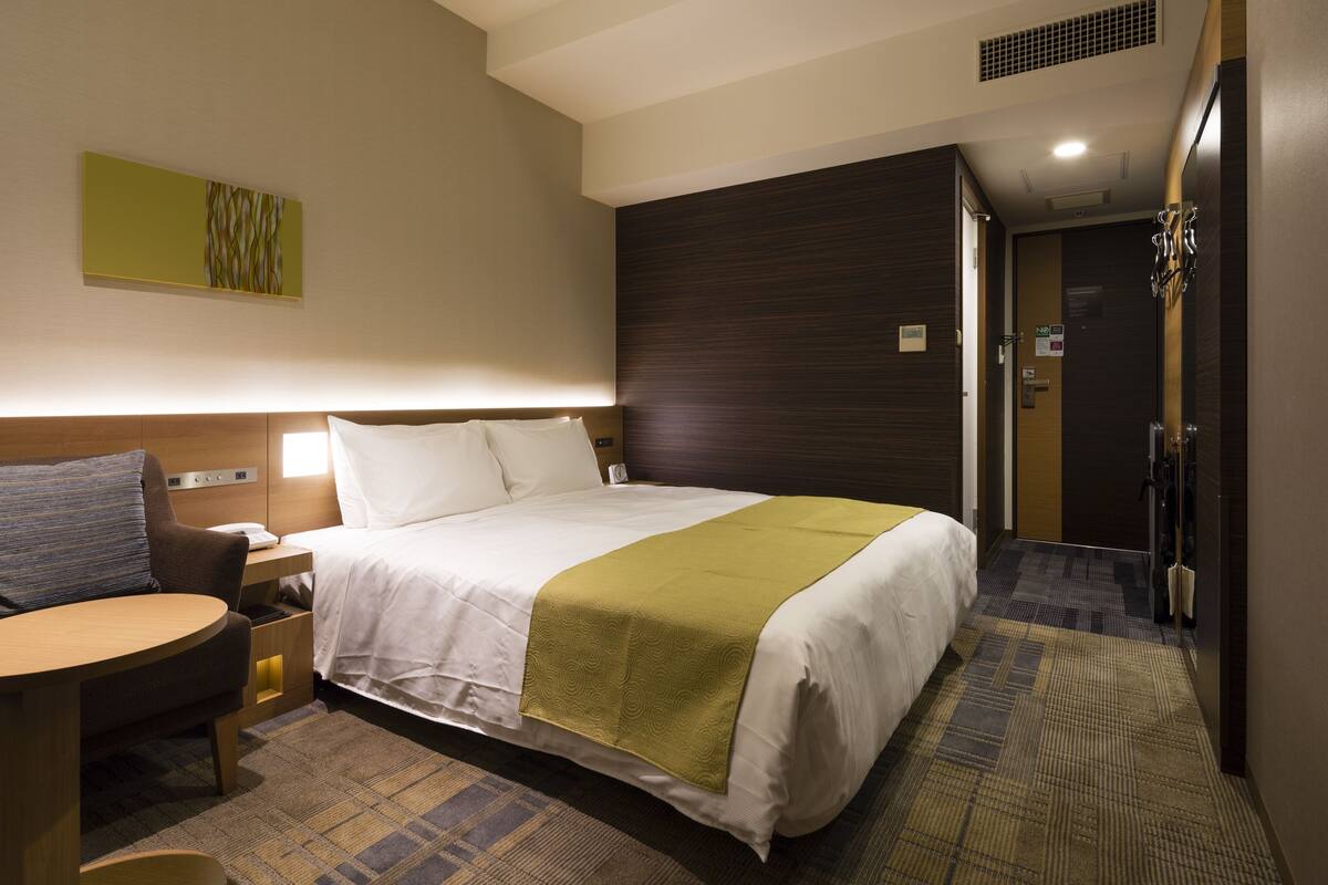 Una habitación de hotel con cama, mesita de noche, silla y una pequeña mesa.