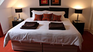 A neatly made bed with a white bedspread, two red and green plaid pillows, and two folded brown towels on the bed. There are two nightstands with lamps, a framed picture on each, and a window with curtains. - The Golden Larches Cafe and B&B (Lochearnhead)