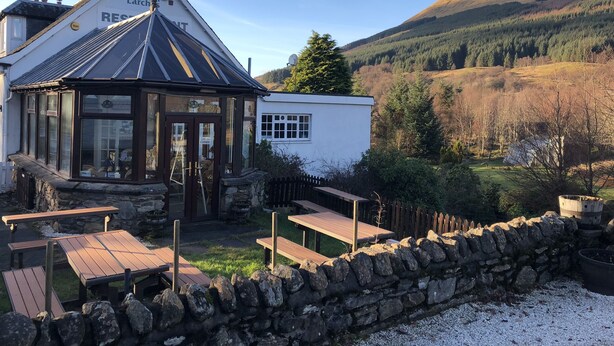 A traditional building with a gabled roof, a stone wall, and wooden benches. - The Golden Larches Cafe and B&B (Lochearnhead)