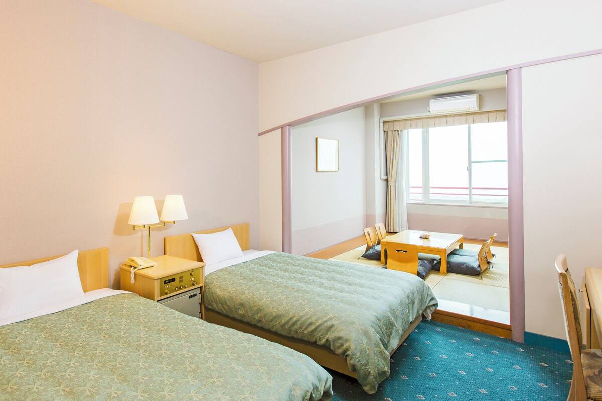 A hotel room with two beds, a desk, a chair, a small table, and a window with curtains.