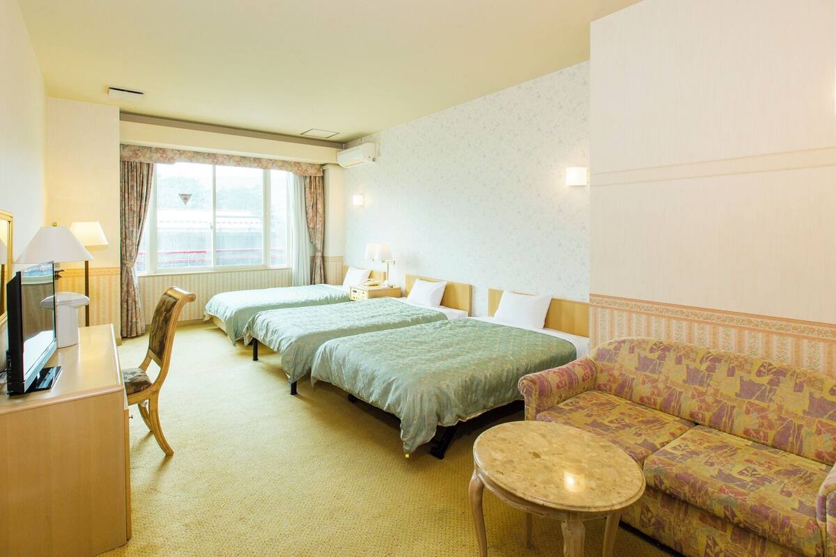A hotel room with two beds, a sofa, a small table, a desk with a lamp, and a window with curtains.
