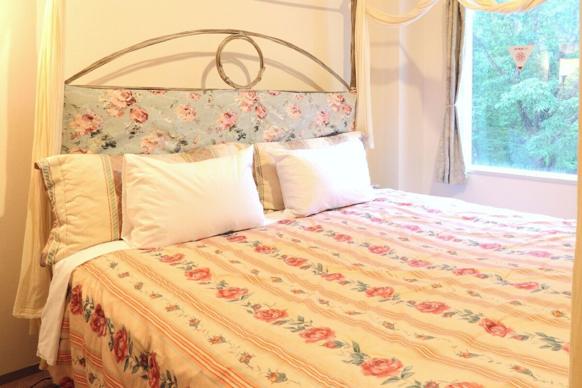 Un lit avec une literie à motifs floraux, une tête de lit en métal et une fenêtre avec des rideaux.