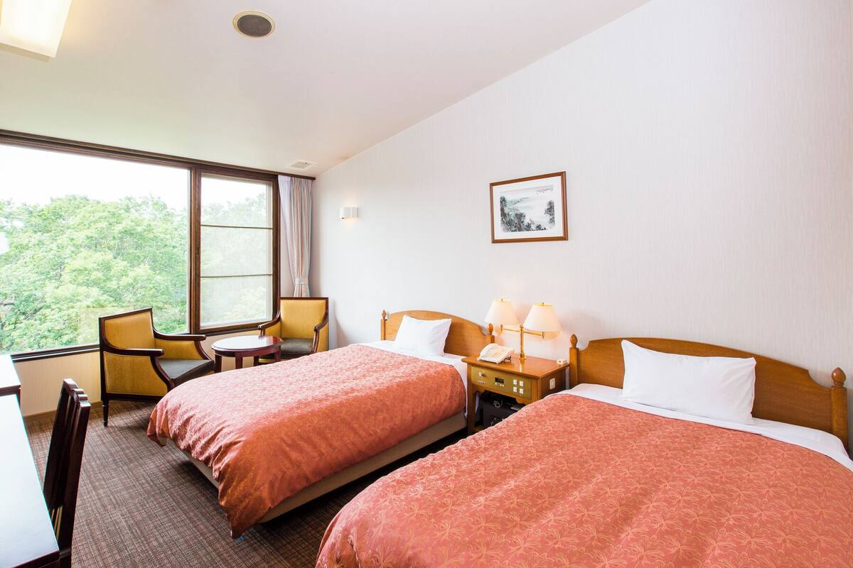 A hotel room with two beds, a desk, a chair, and a window with a view of trees.
