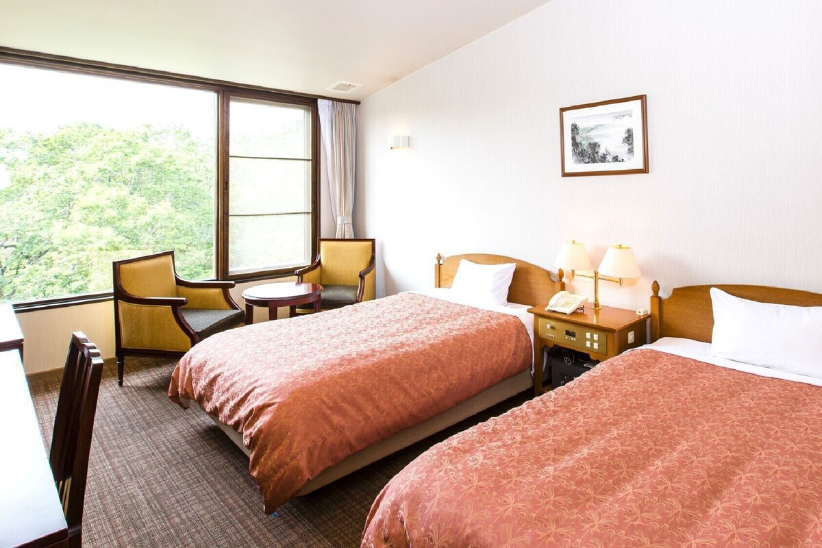 Une chambre d’hôtel avec deux lits, un bureau, des chaises, une fenêtre donnant sur un paysage agréable et un tableau encadré au mur.