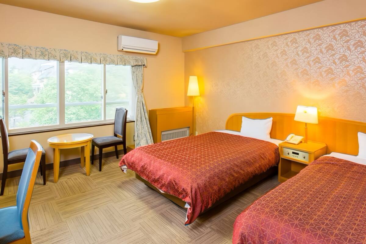 A hotel room with two beds, a small table, a chair, a lamp, and a window with curtains.