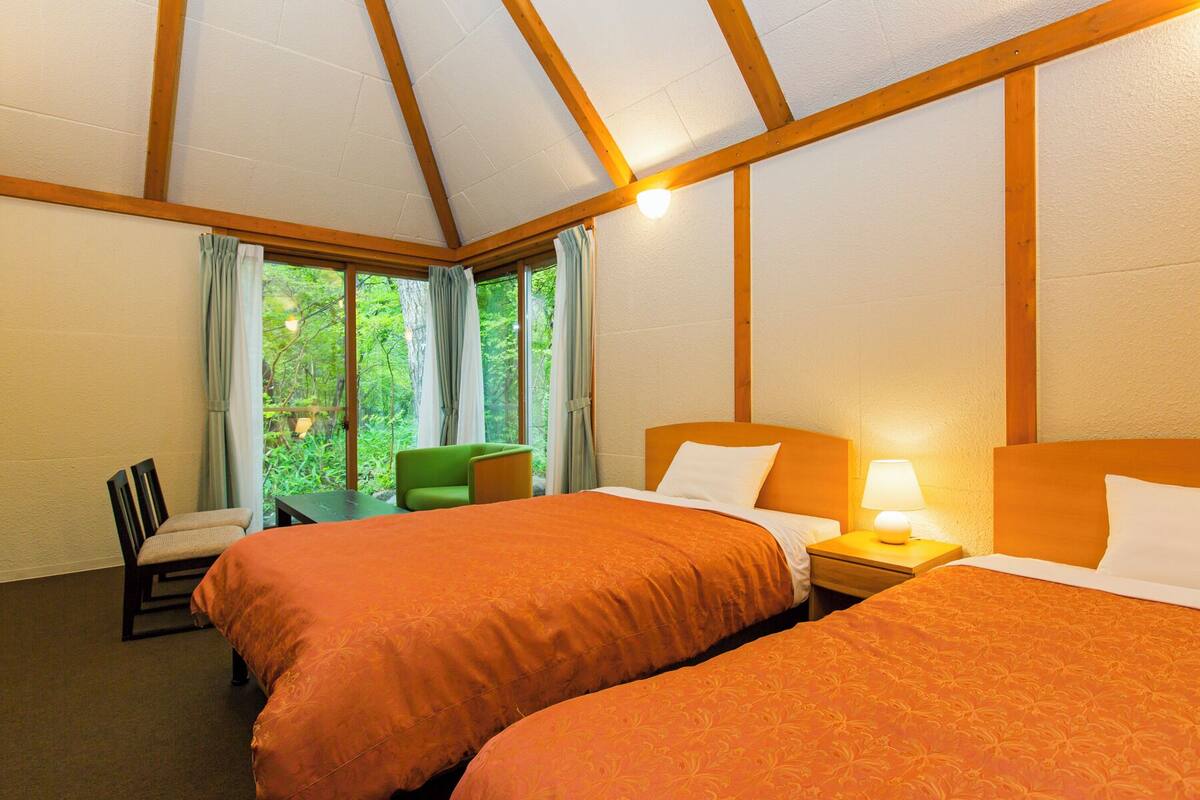 A hotel room with two beds, a desk, and a chair. There are large windows with curtains, and the room has wooden beams on the ceiling.