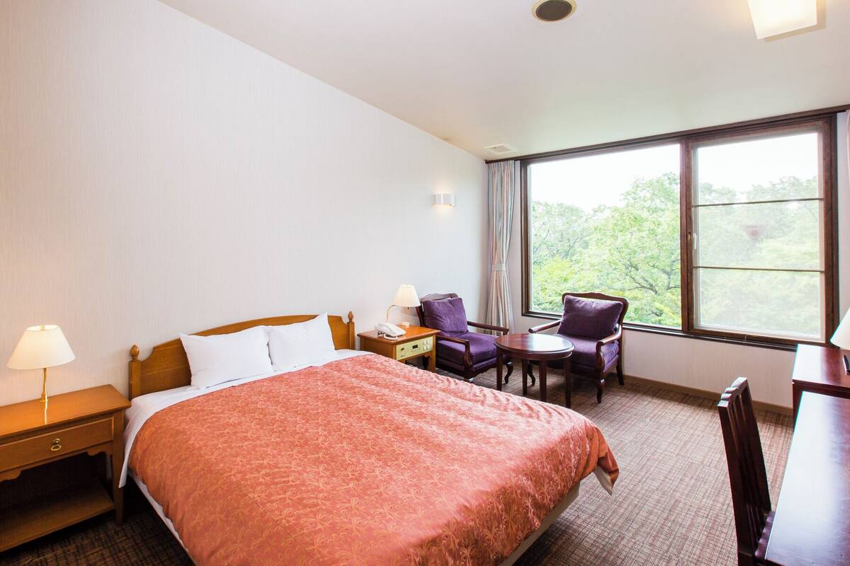 A hotel room with a bed, two chairs, a small table, a nightstand, and a window with a view of trees.