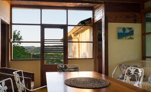 A room with a large window offering a view of greenery and hills. There is a wooden table with a woven placemat and wicker chairs.