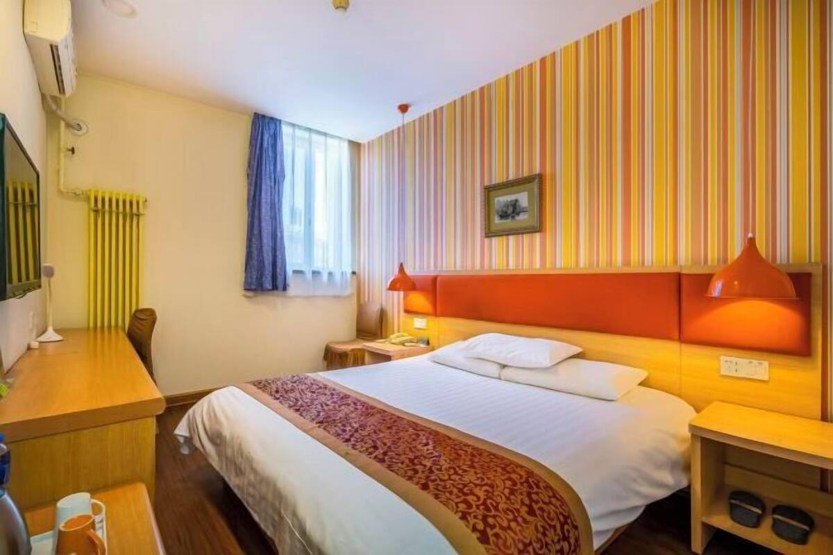 A hotel room with a bed, bedside lamps, a desk, a chair, a radiator, and a window with curtains.