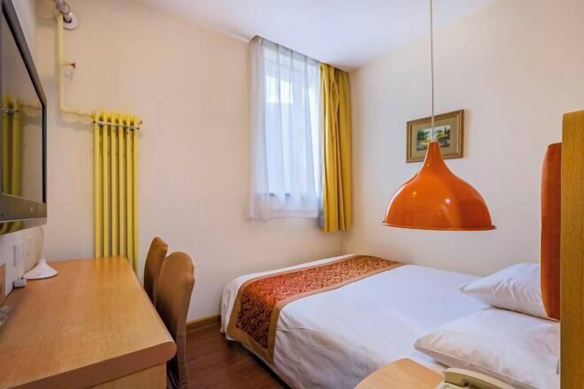 A hotel room with a bed, a desk, and a chair. There is a window with curtains, a painting on the wall, and a hanging lamp.