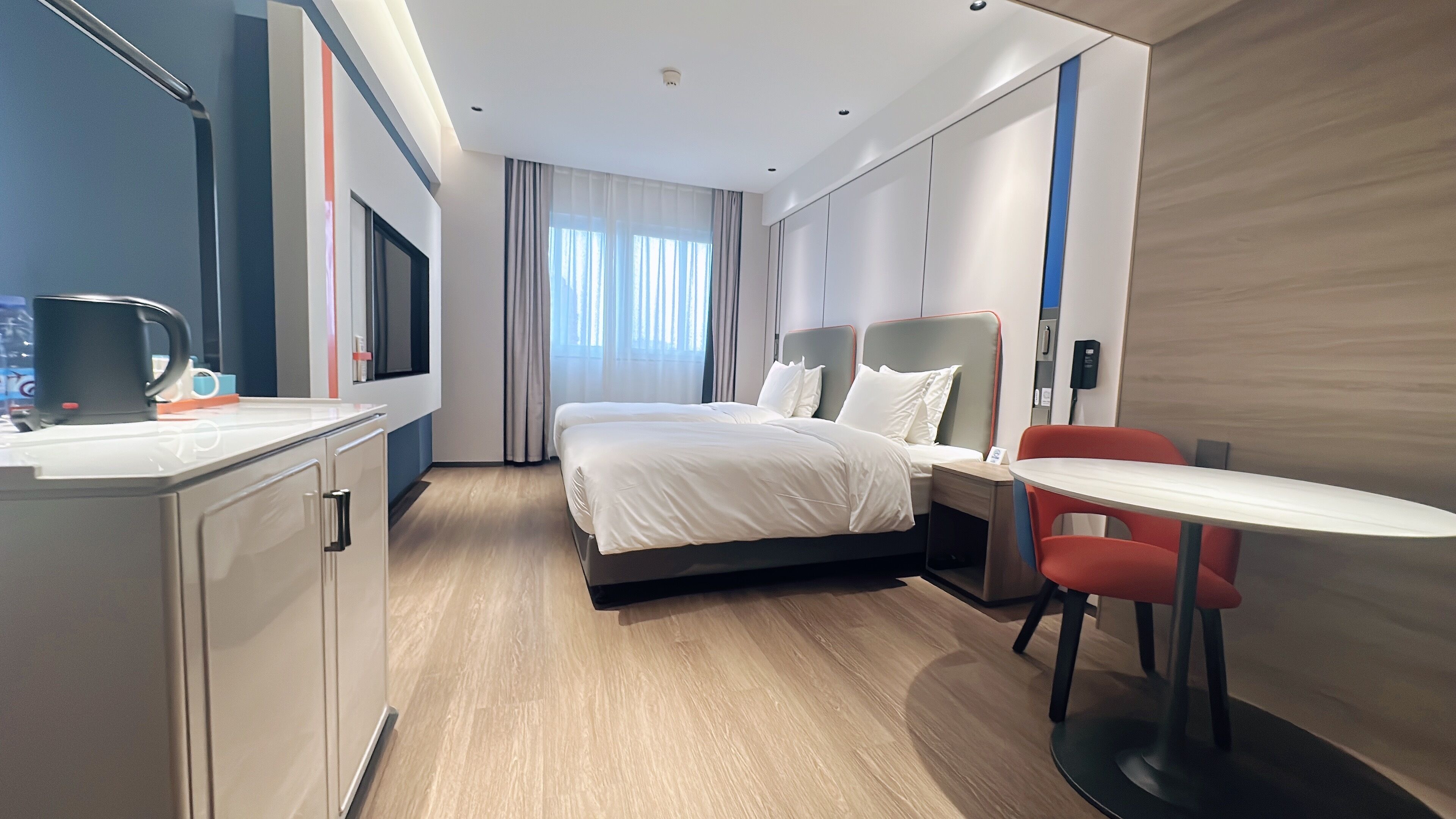A modern hotel room with a bed, bedside table, and a small desk with a chair.