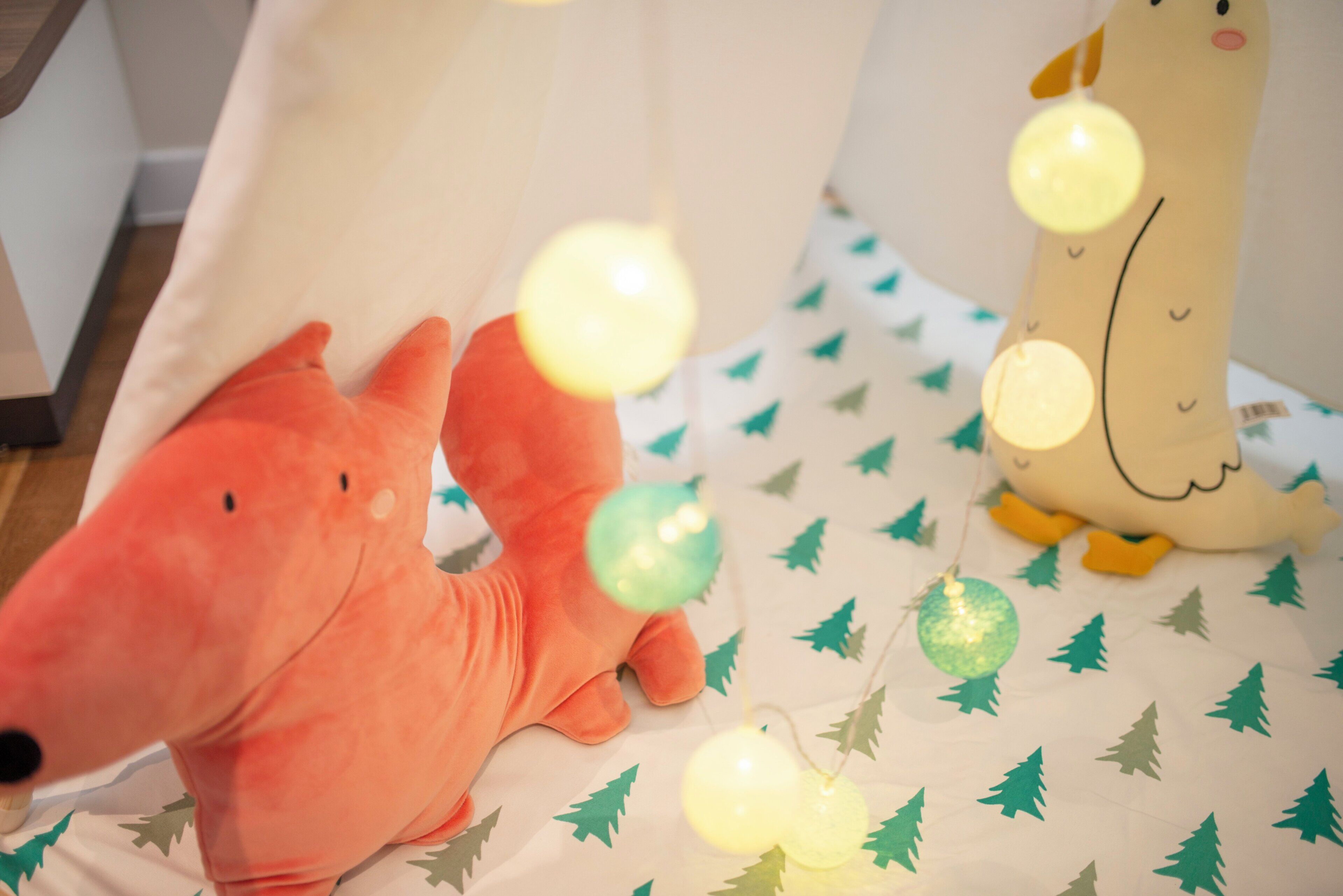 A bed with a white sheet and green tree patterns, a red plush toy, and a brown plush toy.