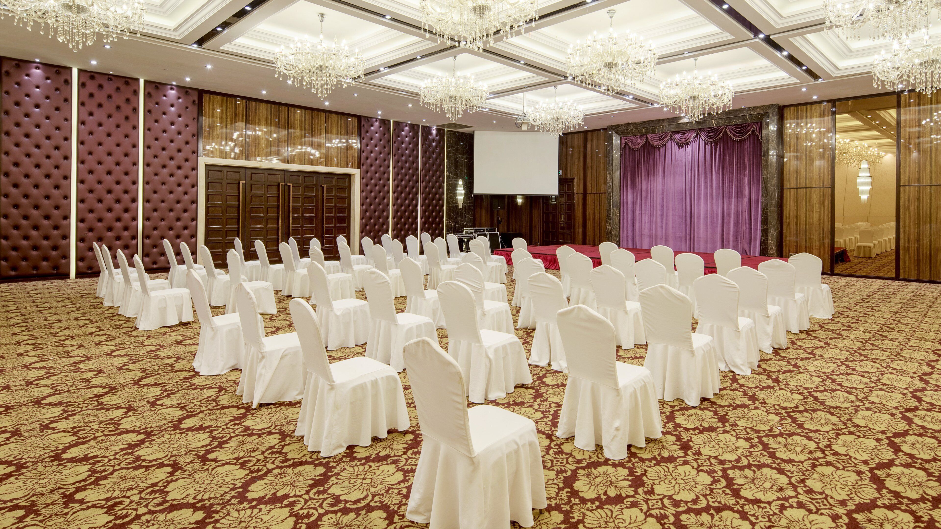 Une salle de conférence avec des rangées de chaises blanches, une scène recouverte d’un tissu rouge et un grand écran.
