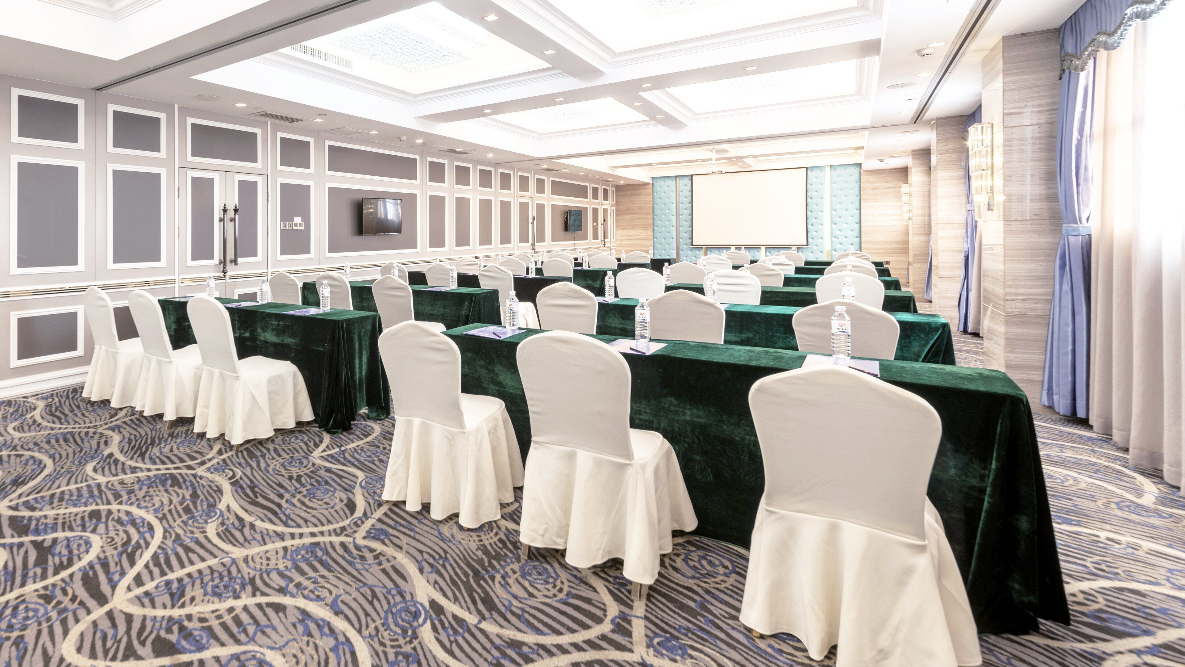 Un salón de conferencias con mesas dispuestas en filas, sillas blancas y una pantalla grande.