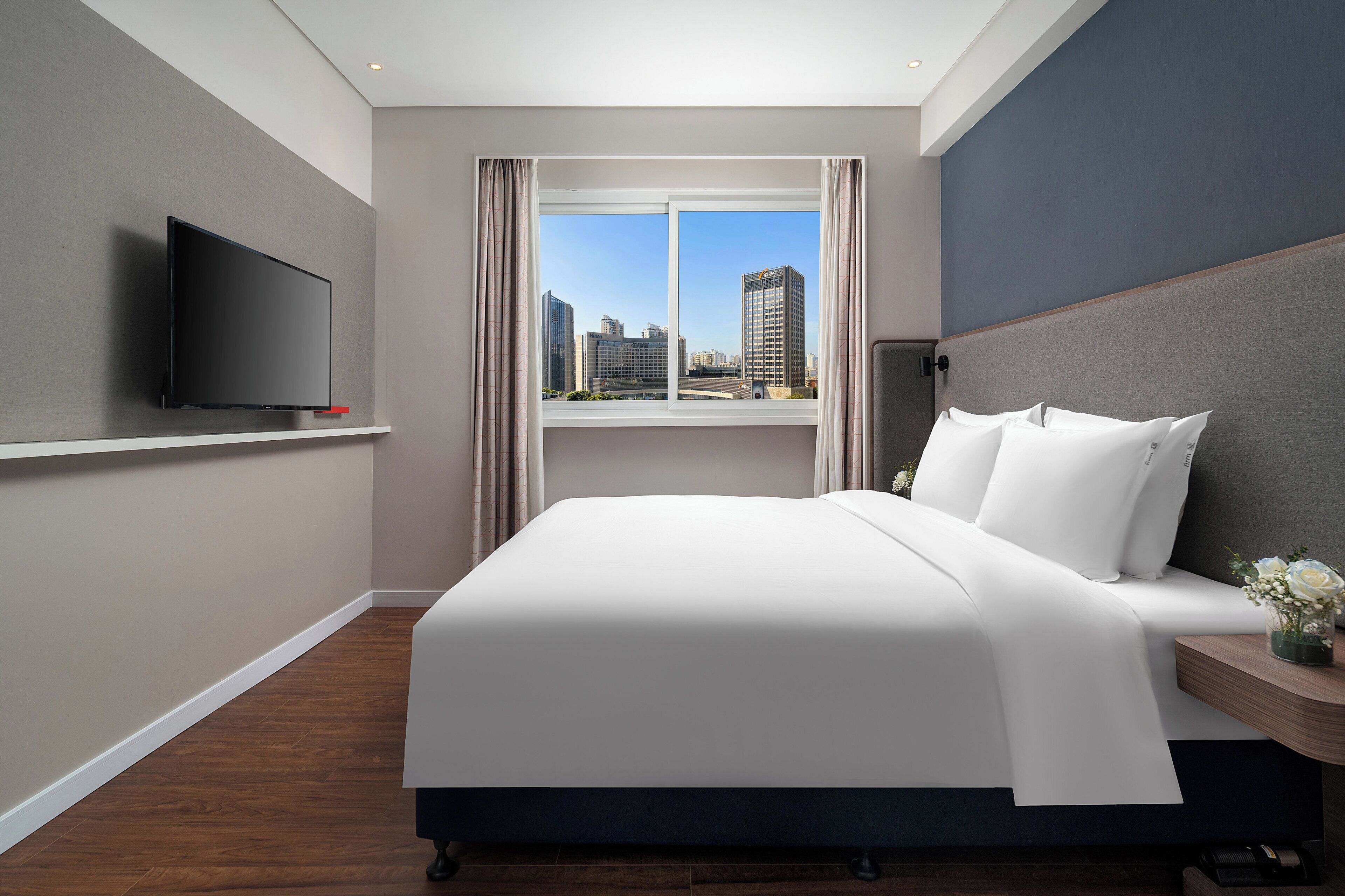 Ein Hotelzimmer mit Bett, Wandfernseher, Fenster mit Stadtblick und Nachttisch mit Blumenvase.