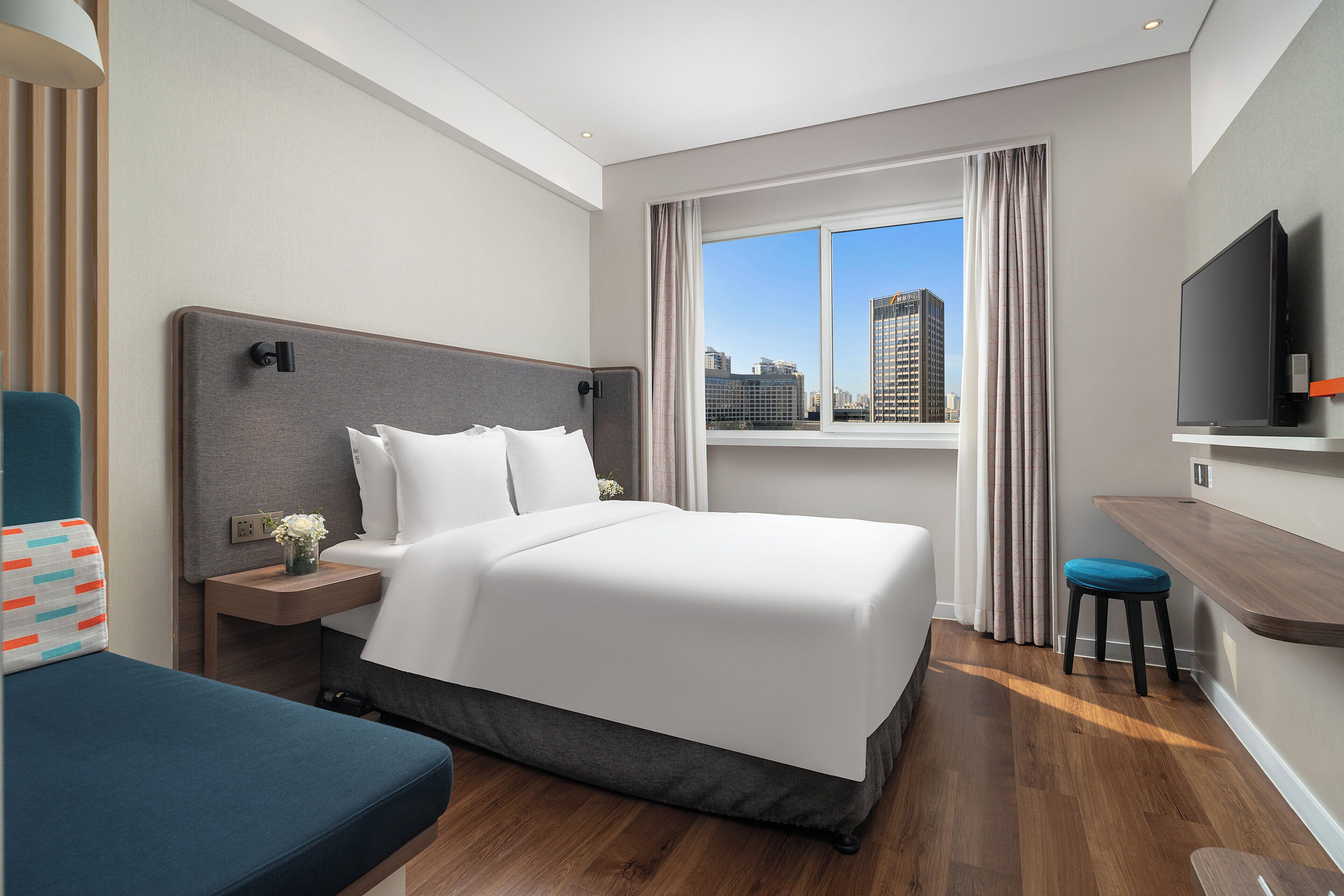 A hotel room with a bed, a desk, a chair, a TV, and a window with a city view.