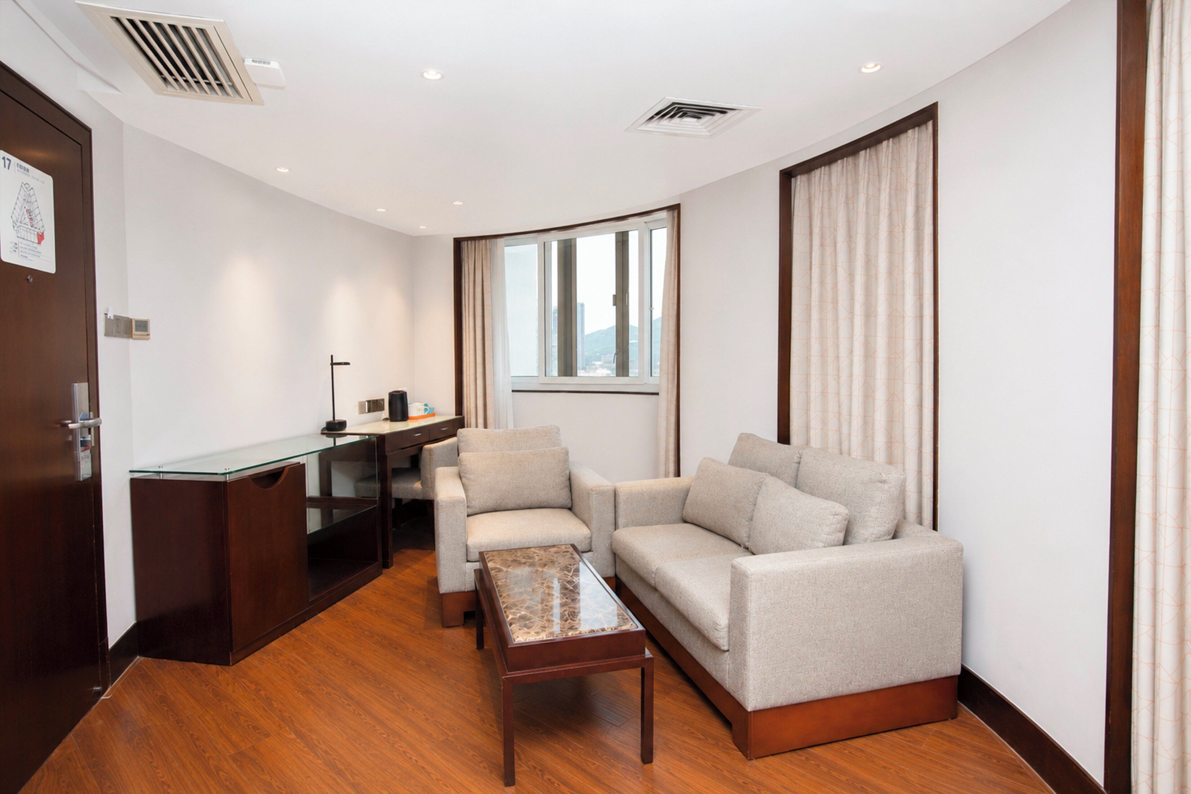 Ein modernes Hotelzimmer mit Holzboden, einer Couch, einem Couchtisch, einem Schreibtisch mit Computer und einem Fenster mit Vorhängen.