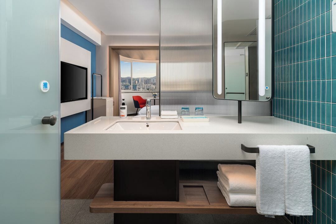A modern bathroom with a large mirror, two sinks, a red chair, and a view of the city through a window.