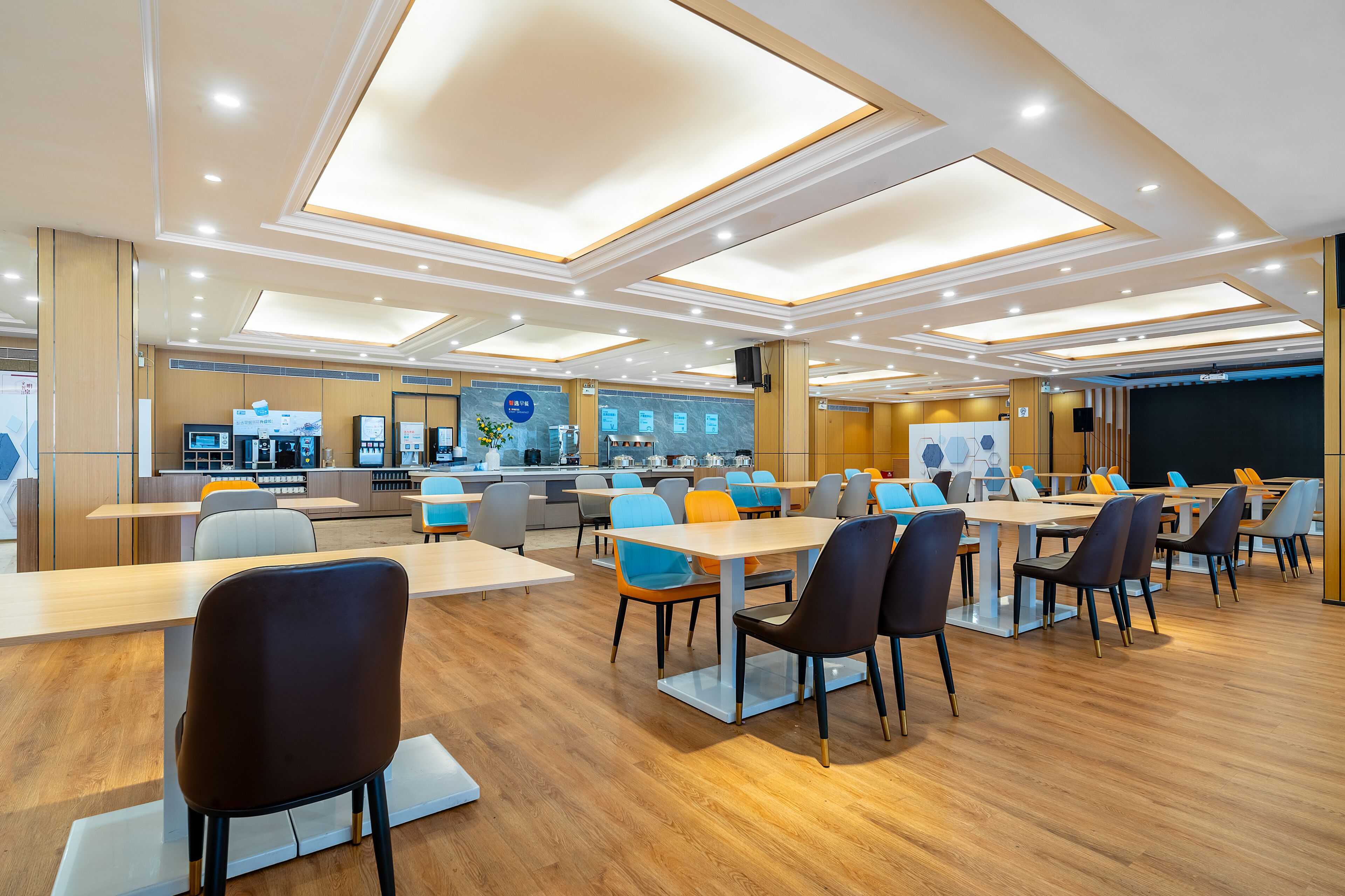 Une salle de conférence moderne, dotée de plusieurs tables et chaises, d’un coin cuisine et d’une scène avec un écran.