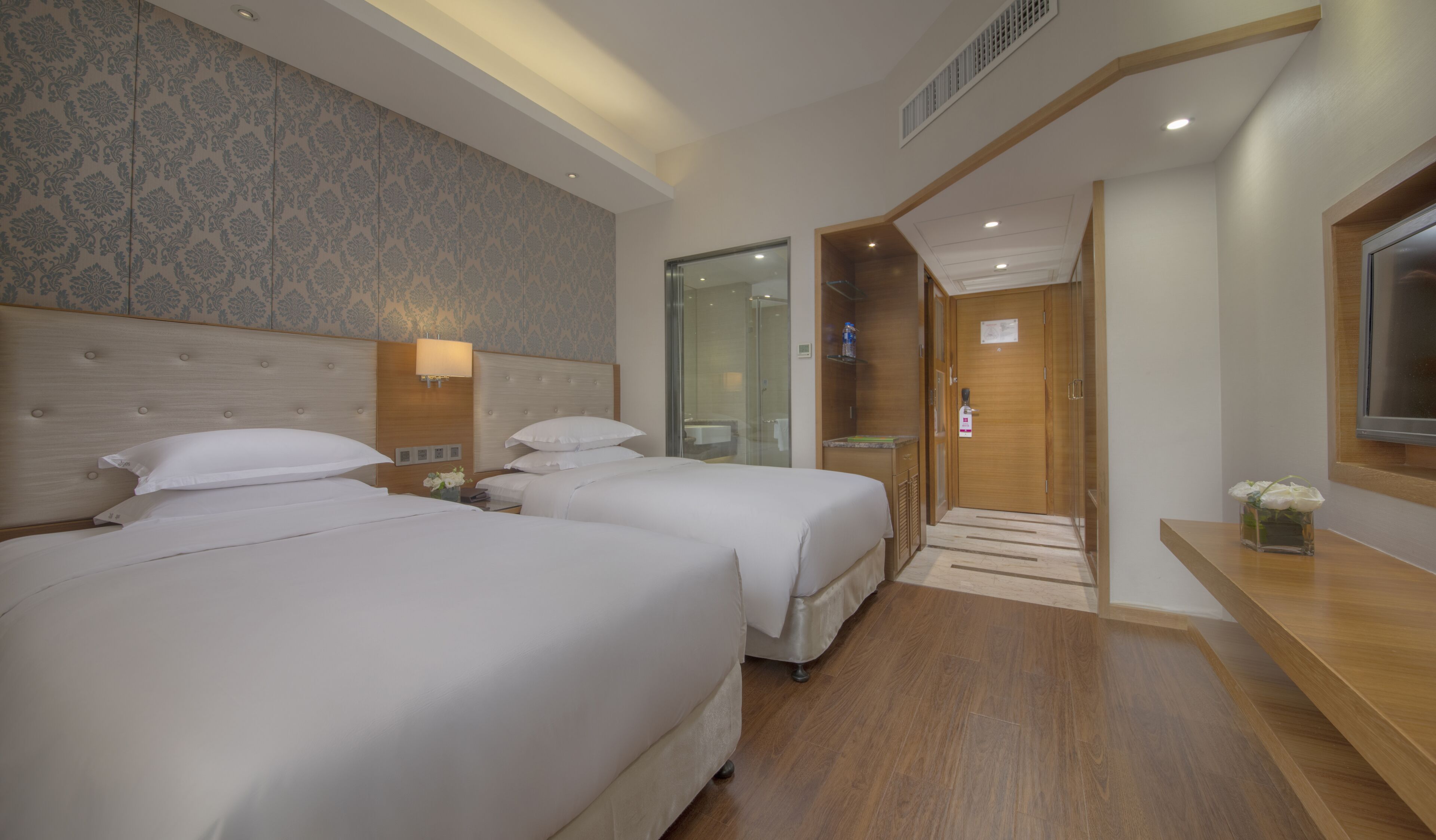 A hotel room with two beds, a wooden headboard, a small bedside table with a lamp, and a shower area.