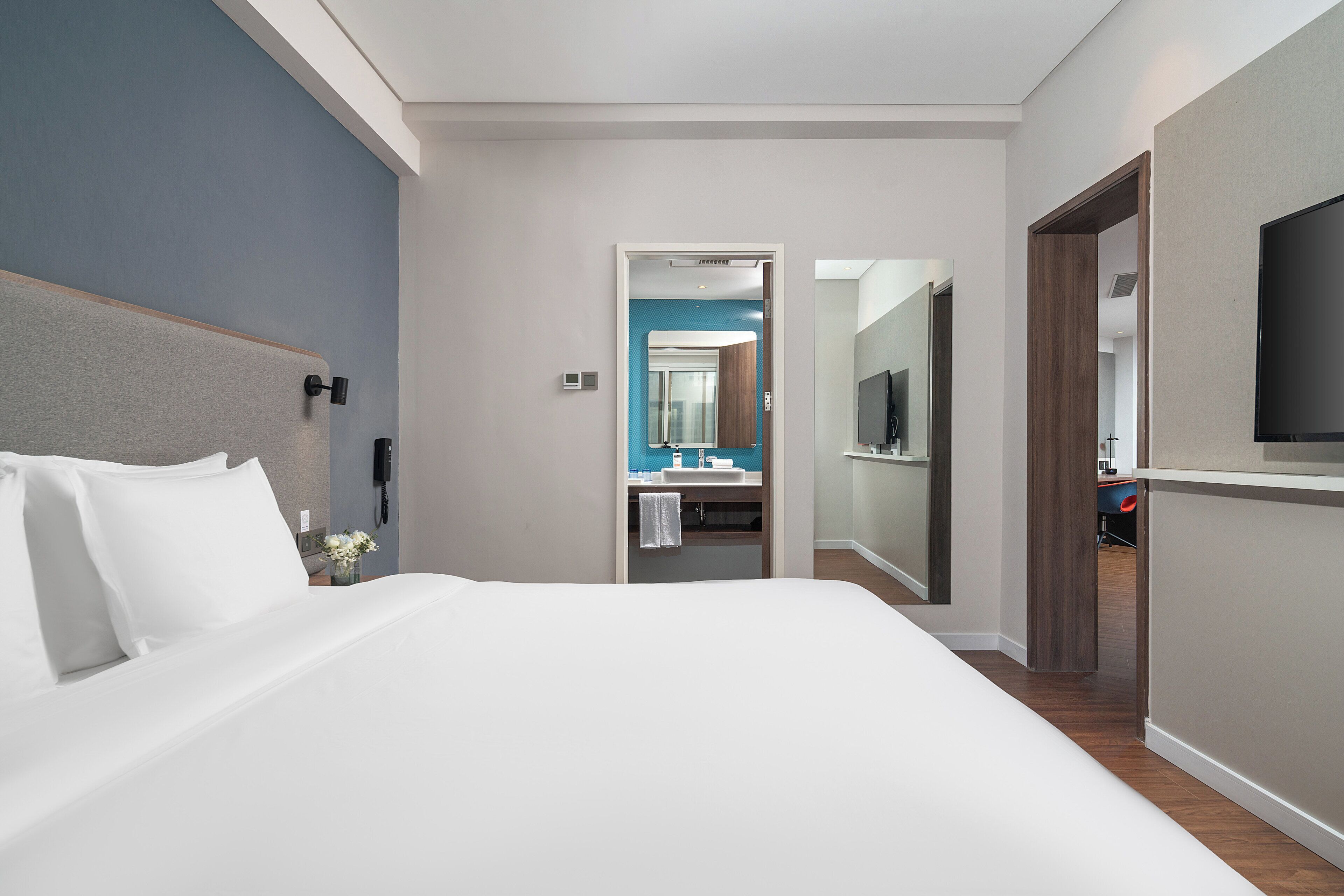 A hotel room with a large bed, a bedside table, a mirror, a TV mounted on the wall, and a bathroom visible through an open door.