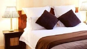 Una cama bien tendida con sábanas blancas y dos almohadones oscuros, flanqueada por dos lámparas de noche en mesitas de madera.