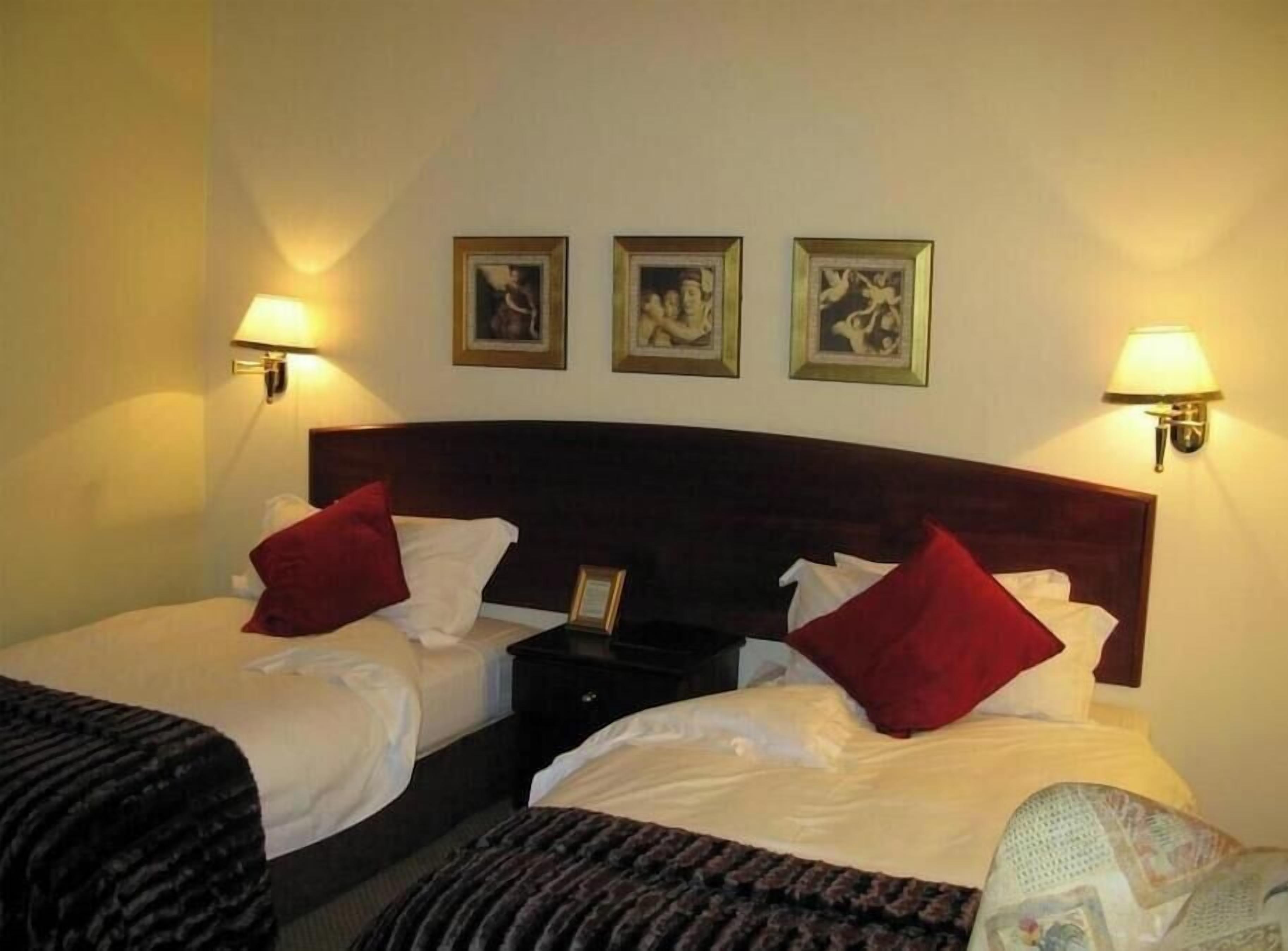 A hotel room with two single beds, a dark wooden headboard, and three framed pictures on the wall.
