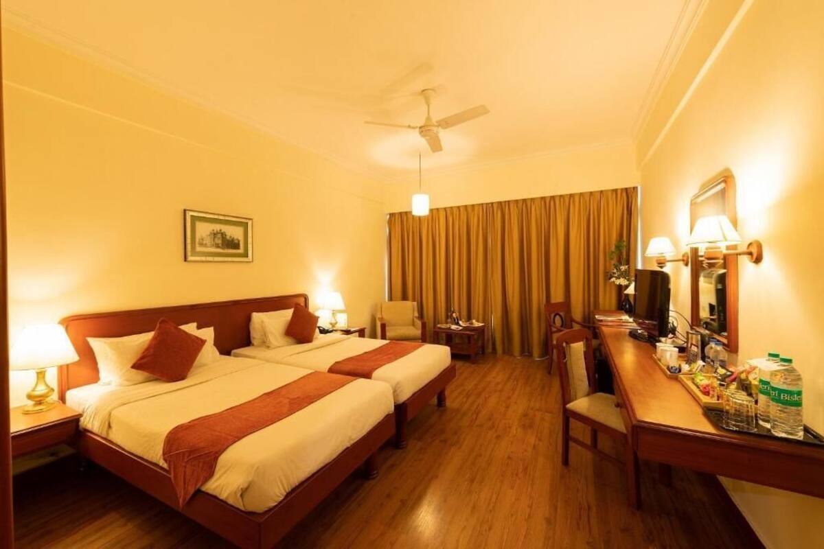 A hotel room with two beds, a desk, a chair, a ceiling fan, and a television.