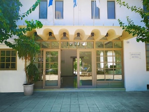 The entrance of Apollon Hotel with a large potted plant, glass doors, and a sign indicating the hotel's name. - Apollon (Agios Nikolaos)