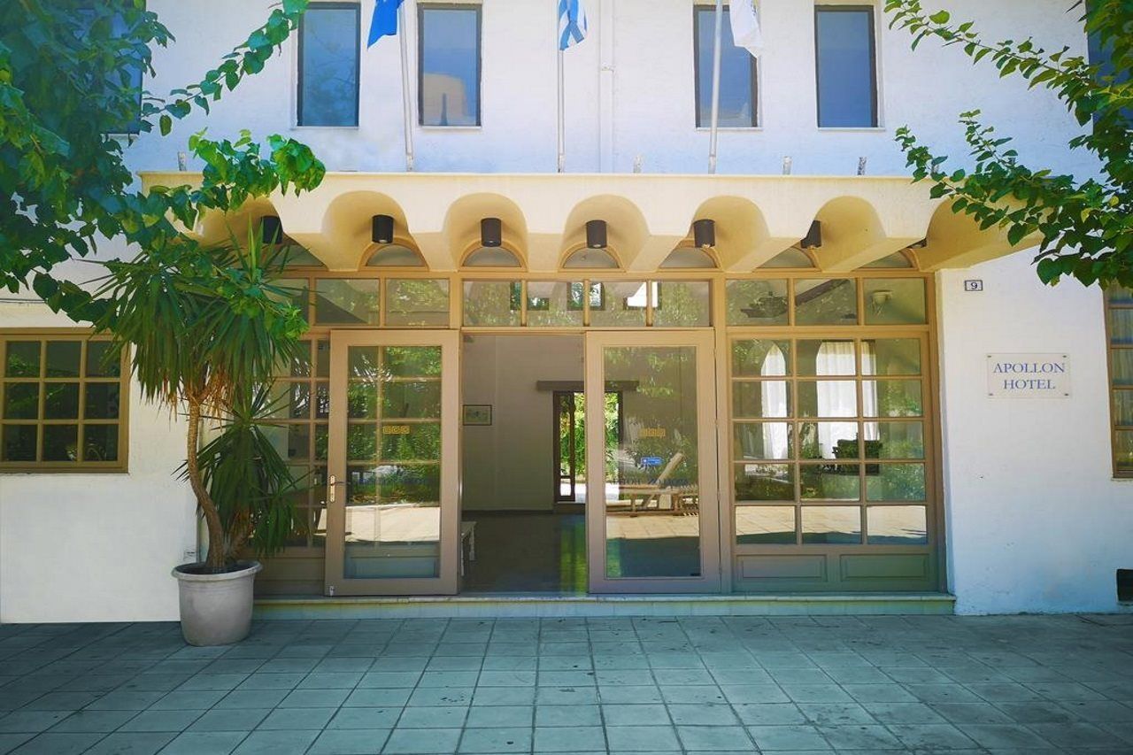 Un edificio con entrada de vidrio, un letrero que dice 'APOLLON HOTEL' y una planta en maceta al frente.