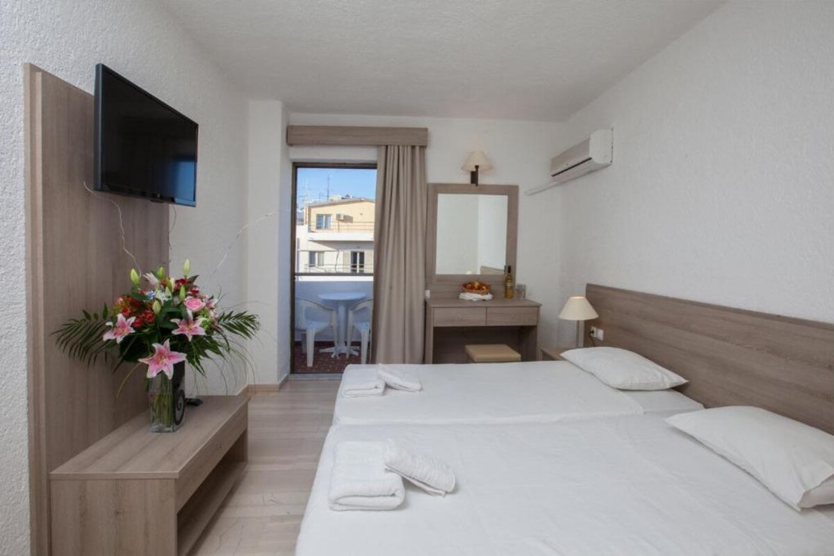 A hotel room with a bed, a TV mounted on the wall, a desk with a lamp, a mirror, and a flower vase.