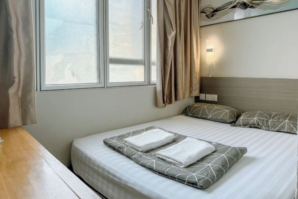 A neatly made bed with pillows and a folded towel, a bedside table, a window with curtains, and a wall-mounted light.