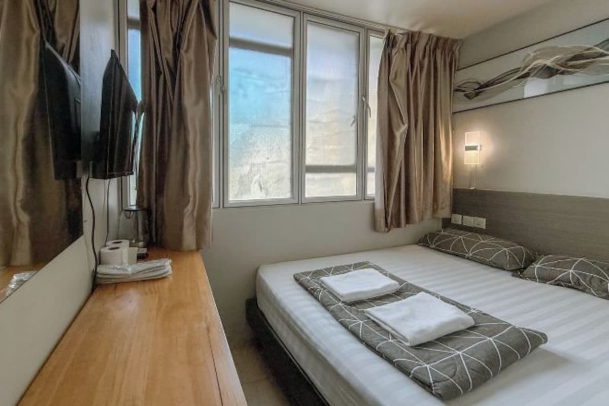 A hotel room with a bed, a bedside table with a TV, a window with curtains, and a wall-mounted light.