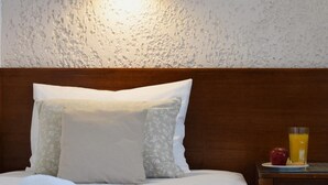 A neatly made bed with a rolled towel, a bedside table with a glass of orange juice and an apple, and a wall-mounted light fixture. - Altis Hotel (Malia)