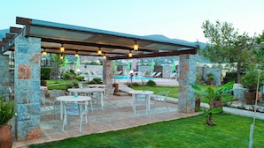 A covered outdoor seating area with stone pillars, tables, and chairs, overlooking a pool and greenery. - Altis Hotel (Malia)
