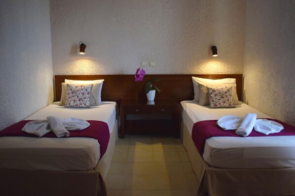 A hotel room with two beds, each with floral pillows, a nightstand with a potted plant, and wall-mounted lights. - Altis Hotel (Malia)