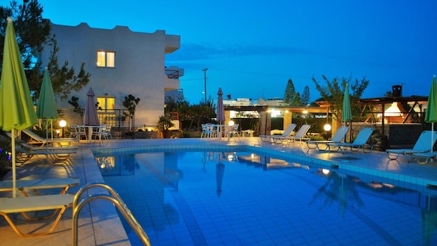 A swimming pool with lounge chairs and umbrellas, a building with a balcony, and a view of the sea at dusk. - Altis Hotel (Malia)