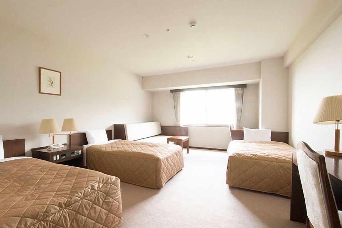 A hotel room with two single beds, a desk with a lamp, a chair, a small table, and a window with curtains.