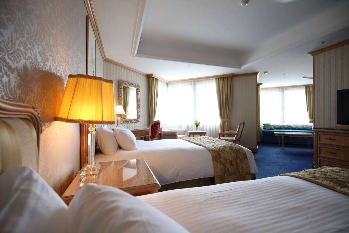 Habitación de hotel con dos camas, zona de estar con un sofá y sillones, televisor y un ventanal grande con cortinas.