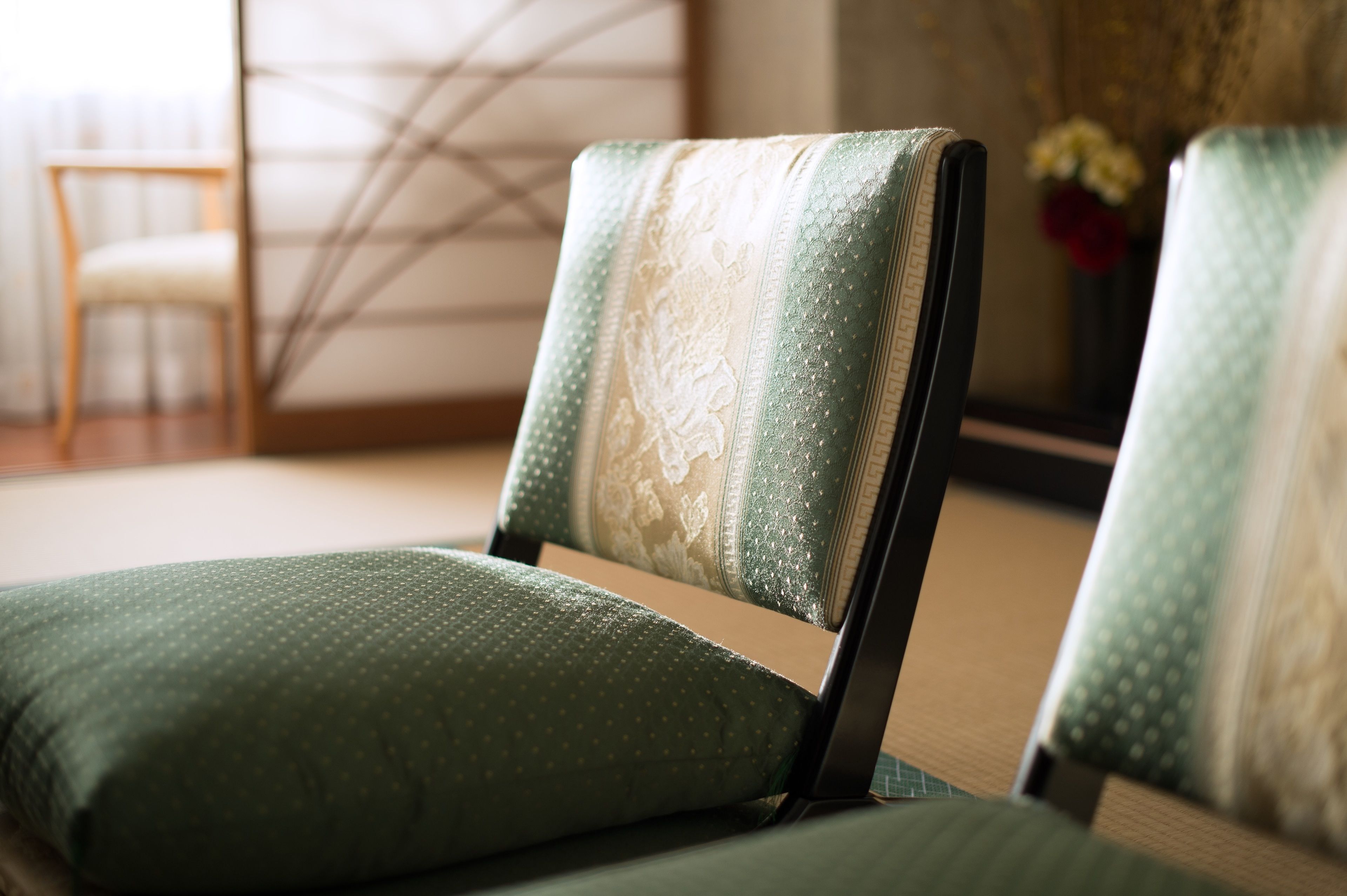 A room with a green upholstered chair and a matching cushion, a wooden chair in the background, and a vase with flowers on a table.