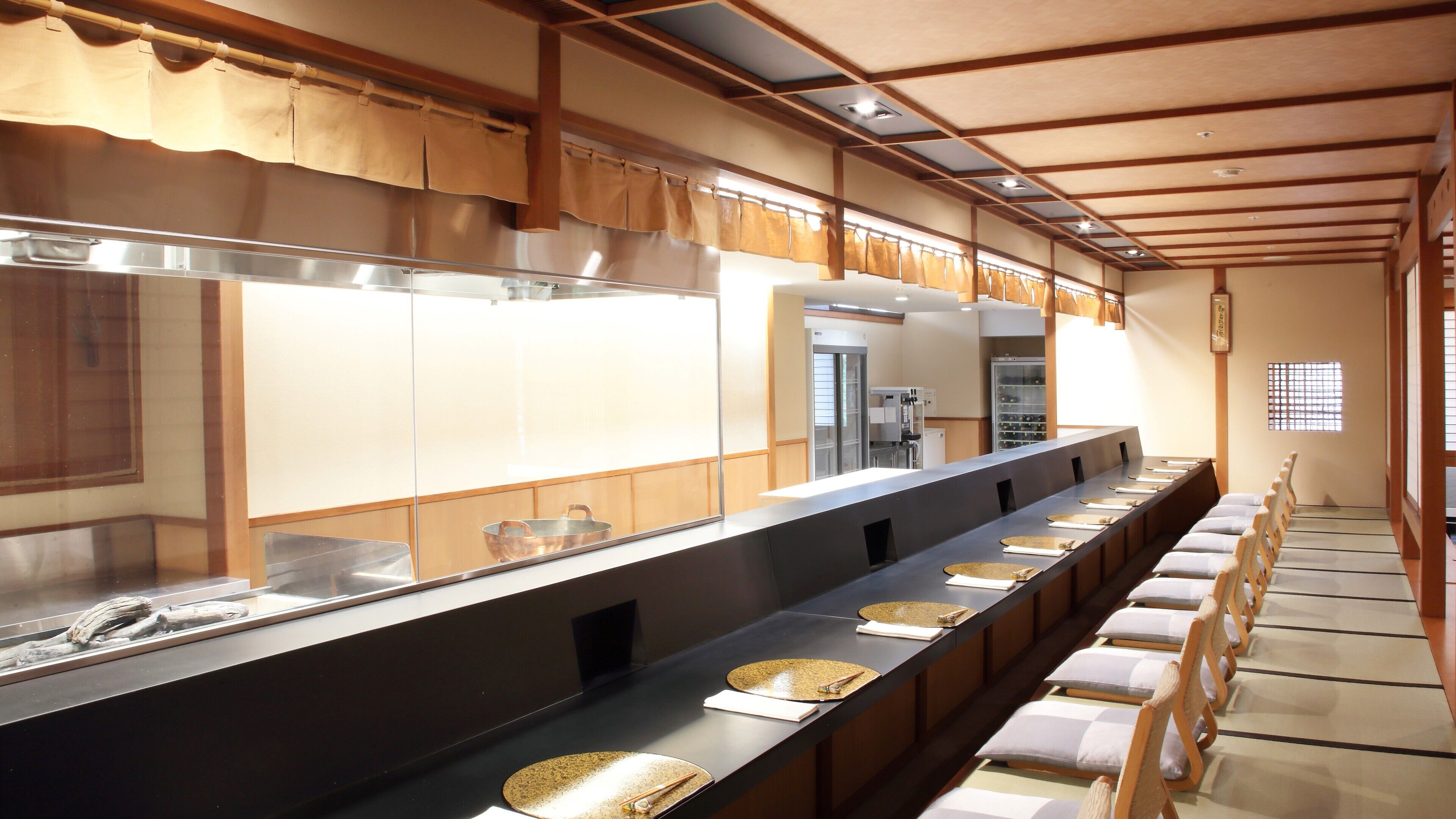 A long, dark counter with multiple round, gold-topped plates and chopsticks, flanked by wooden chairs with cushions.